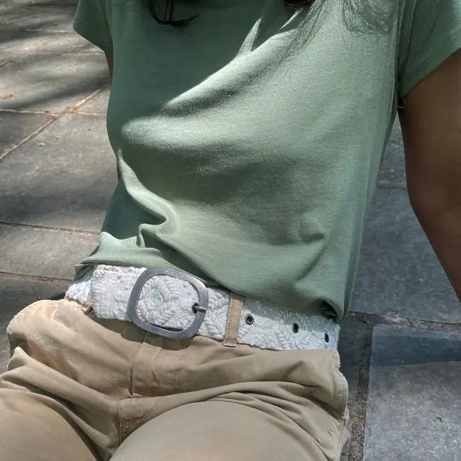 A person sits outdoors on stone pavement, shown from torso to lap, wearing a light green t-shirt, beige pants, and the JENNY KRAUSS - WANDERING CLOUDS EMBROIDERED BELT with a decorative buckle. Head and lower legs are not visible.