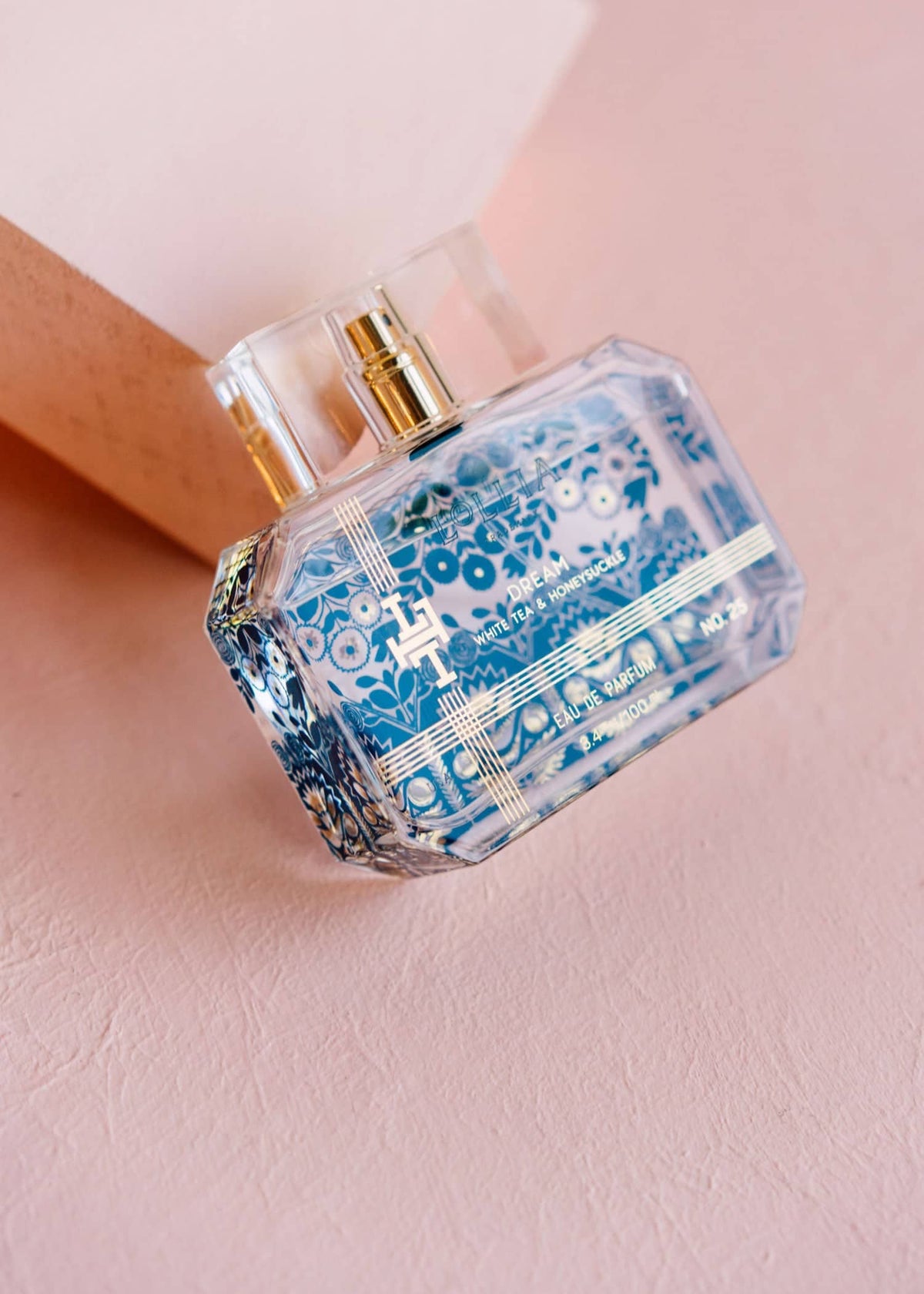 A rectangular perfume bottle with intricate blue patterns and gold accents lays on a pink surface, leaning against a light pink block. The elegant bottle, labeled DREAM EAU DE PARFUM by MARGOT ELENA, features a clear cap that allows visibility of the liquid inside.