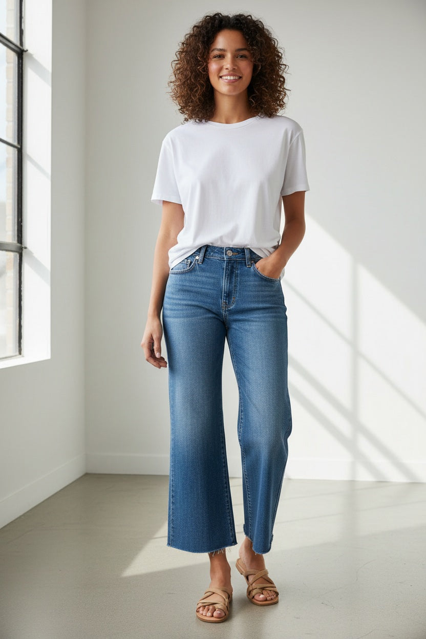 A person stands outdoors on grass, wearing the FRANK AND EILEEN - GALWAY WIDE LEG JEAN IN 2002 MID WASH and a white shirt, with their right hand relaxed by their side.