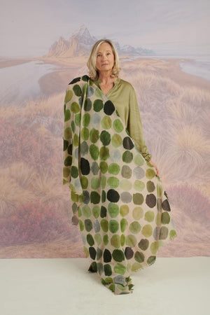 A woman with blonde hair displays the STORIATIPIC - BETTY WOOL SCARF, a large rectangular wool accessory with earth-toned polka dots, set against a scenic mountain landscape.