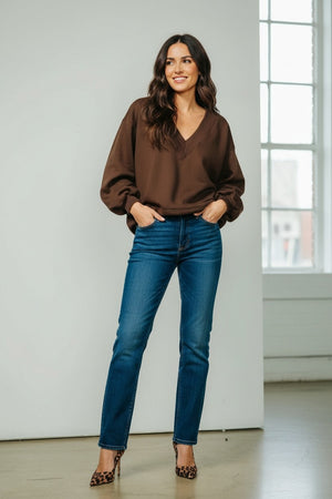 A woman stands indoors, smiling with her hands in her pockets, wearing the KMJ Tucker V Neck Sweater in Chocolate by KMJ—a relaxed fit brown long-sleeve top—paired with blue jeans and leopard print heels. A large window is visible in the background.