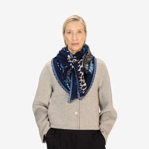A woman with light hair pulled back wears a light gray jacket and the INOUI EDITIONS PAPILLON (BUTTERFLIES) square wool scarf in blue by INOUI. She stands against a plain white background, looking straight at the camera with her hands in her pockets.