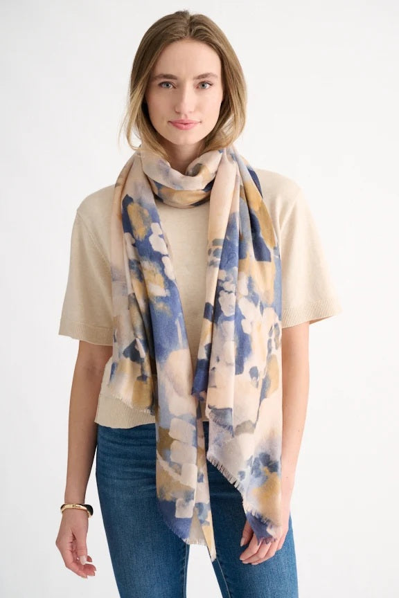 A person wearing a beige short-sleeve top and blue jeans stands against a plain white background, styled with the KINROSS CASHMERE Abstract Print Scarf in Navy—a luxurious accessory by KINROSS woven from a sumptuous blend of wool, silk, and cashmere in softly patterned blue, beige, and gold tones.