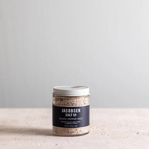 A small glass jar of JACOBSEN SALT CO. JACOBSEN SALT - INFUSED BLACK PEPPER SALT with a white lid rests on a light beige surface against a light gray background. The dark label with white text highlights this floral, spicy blend.