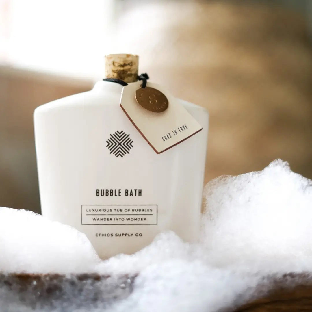 A vegan bubble bath in a luxurious white bottle from Ethics Supply Co features a cork stopper and tag. The label, "Soak In Love Ceramic Bubble Bath," offers an enchanting experience with its promise of abundant bubbles.