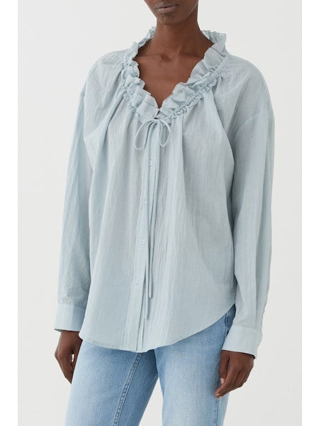 A person wears the ELEH RUFFLE NECK BLOUSE in light blue cotton with a ruffled collar, V-neck, and front buttons, paired with light blue jeans, standing against a plain white background.