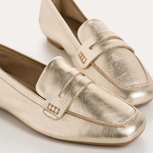 The REQINS - HEMA Metallic Loafer in Platinum features shimmering leather, a low heel, and a decorative front strap, showcased on a plain white background.