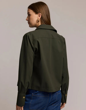 A woman with long brown hair models the HINSON WU - ASTRID LONG SLEEVE TRAVELER'S TWILL SHIRT JACKET in dark green over an olive top and blue jeans, showcasing versatile HINSON WU travel style with gold hoop earrings against a neutral background.