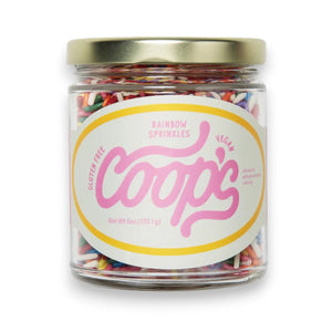 A glass bowl of ice cream topped with COOP’S - RAINBOW SPRINKLES, caramel sauce, and a cherry sits next to an open jar of Coop’s Salted Caramel Sauce and bowls of toppings on a table.