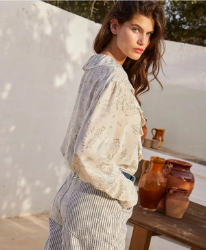 A woman with long brown hair stands outdoors, wearing the MOMONI RUFFLED BLOUSE and striped pants. She looks over her shoulder, with clay jugs and a wooden table in the background against a white wall.