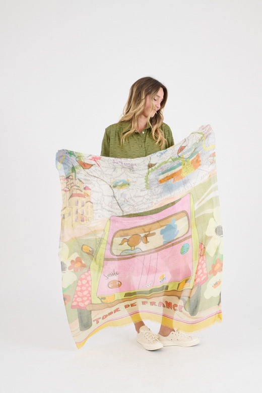 A woman in a green dress drapes the STORIATIPIC Gabie Scarf in Rose over her shoulders. This luxury silk accessory by STORIATIPIC features playful illustrations, maps, and "TOUR DE FRANCE" on a light background.