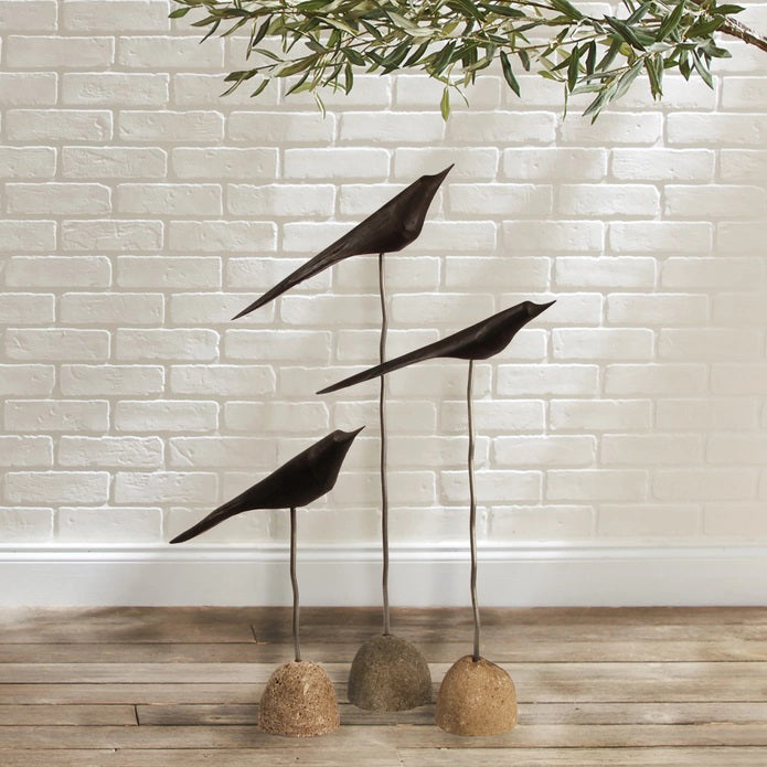The NAPA HOME AND GARDEN &quot;THE FLOCK&quot; Set of 3-Black minimalist bird sculptures are displayed indoors on slender stands with stone bases, set on a wooden floor against a white brick wall with leafy branches above.