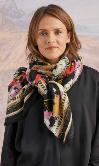 A woman outdoors with a blurred mountain backdrop wears a cream button-up and the STORIATIPIC DORIS Wool Silk Scarf by STORIATIPIC, featuring green, navy, and yellow patterns in a luxe wool-silk blend.