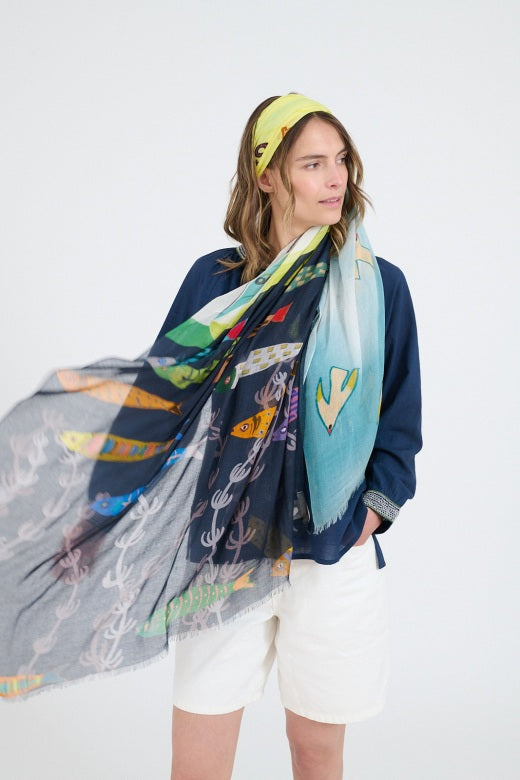 A woman with shoulder-length wavy hair models the STORIATIPIC - GERTRUD SCARF, styled around her neck over a navy blue top, against a plain white background.