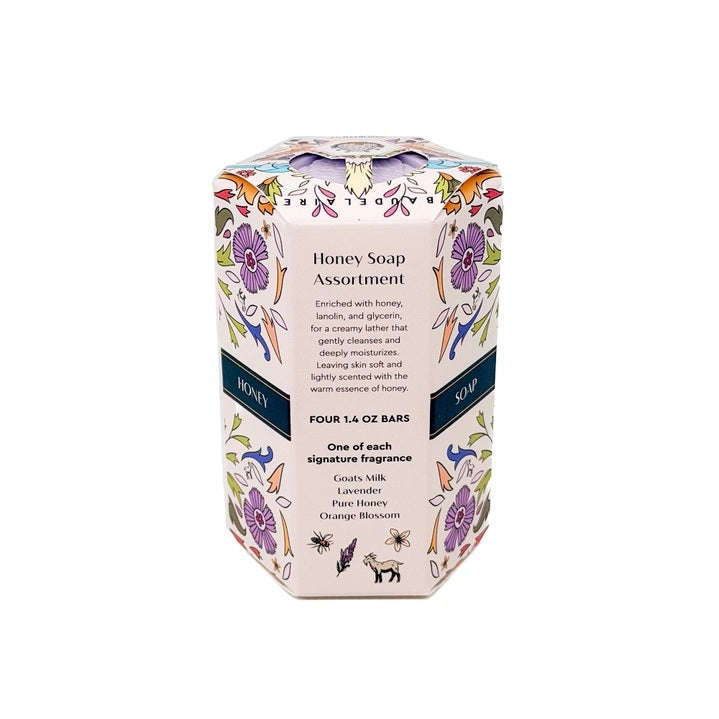 Four pastel-colored, triple-milled soaps—lavender, pink, white, and orange—are stacked next to a decorative floral-patterned box labeled “BAUDELAIRE - HONEY SOAP ASSORTMENT 1.4 OZ QUARTET” by BAUDELAIRE.