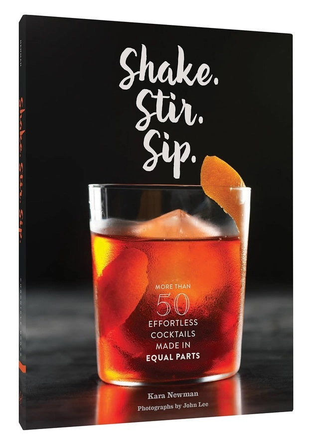 The SHAKE. STIR. SIP. BY KARA NEWMAN recipe book from CHRONICLE BOOKS rests on a light marble surface alongside an orange cocktail with ice, a metal jigger, and a cocktail strainer.