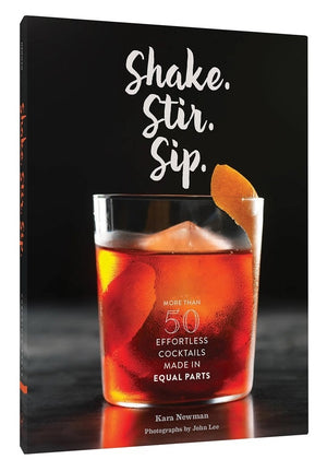 The SHAKE. STIR. SIP. BY KARA NEWMAN recipe book from CHRONICLE BOOKS rests on a light marble surface alongside an orange cocktail with ice, a metal jigger, and a cocktail strainer.