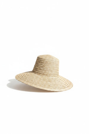Wearing the MERSEA MOROCCAN RUSTIC STRAW HAT, a woman in an orange top and white shorts smiles on a swing, surrounded by plants and hanging lights, enjoying a bright sunny day.