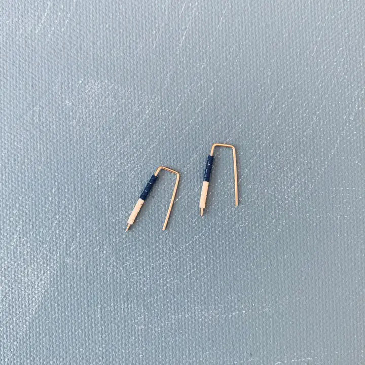 The ALICE RISE LITTLE QUILL EARRINGS IN NAVY AND CHALK are two small, L-shaped gold filled earrings with deep navy centers, shown parallel on a textured gray surface with their tips pointing downward.