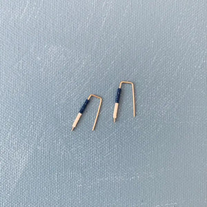 The ALICE RISE LITTLE QUILL EARRINGS IN NAVY AND CHALK are two small, L-shaped gold filled earrings with deep navy centers, shown parallel on a textured gray surface with their tips pointing downward.
