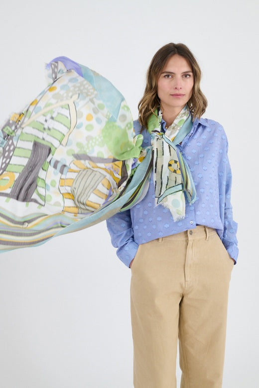 A woman stands against a white background, wearing a light blue textured blouse, beige pants, and the STORIATIPIC - HEIDI SCARF—a colorful cotton accessory from STORIATIPIC—embodying effortless casual elegance.