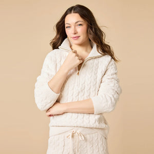 A woman wearing a marshmallow-soft, cream-colored SOFTIES - CABLE KNIT MARSHMALLOW ½ ZIP WITH TAPERED PANT SET, featuring a half-zip pullover and matching pants with a drawstring waist.