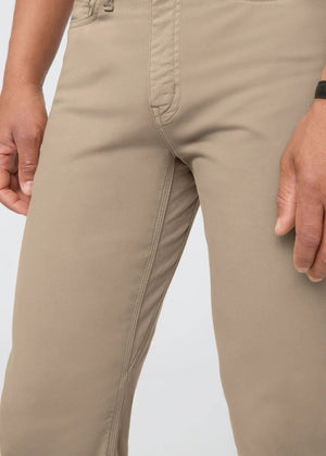 A man with short dark hair stands against a white background, wearing DUER No Sweat Straight Pants in Desert Khaki, a white t-shirt, and white sneakers. He faces the camera with a neutral expression.
