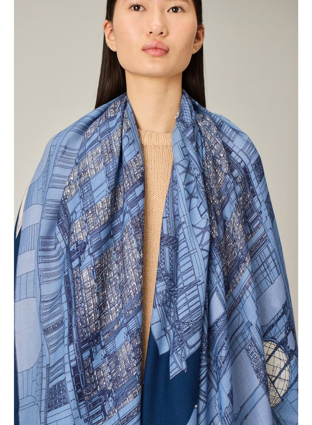 A woman stands against a plain background, wearing a beige sweater, cream wide-leg pants, black shoes, and the INOUI EDITIONS - VIVIENNE WOOL SCARF IN BLUE by INOUI—a long blue scarf with a white pattern made from 100% wool in India and measuring 100 x 190 cm.