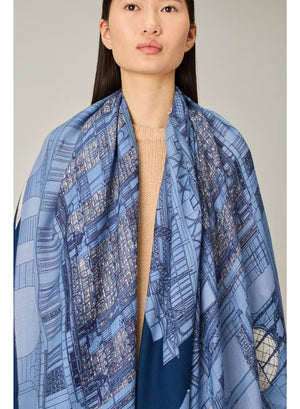 A woman stands against a plain background, wearing a beige sweater, cream wide-leg pants, black shoes, and the INOUI EDITIONS - VIVIENNE WOOL SCARF IN BLUE by INOUI—a long blue scarf with a white pattern made from 100% wool in India and measuring 100 x 190 cm.