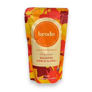 A vibrant package of BRODO BROTH COMPANY’s BRODO BROTH- SPICY NOONA ROASTED GARLIC & CHILI BONE BROTH ORGANIC rests on a wooden surface, surrounded by dried chili peppers, roasted garlic, and a steaming cup of flavorful chicken bone broth.