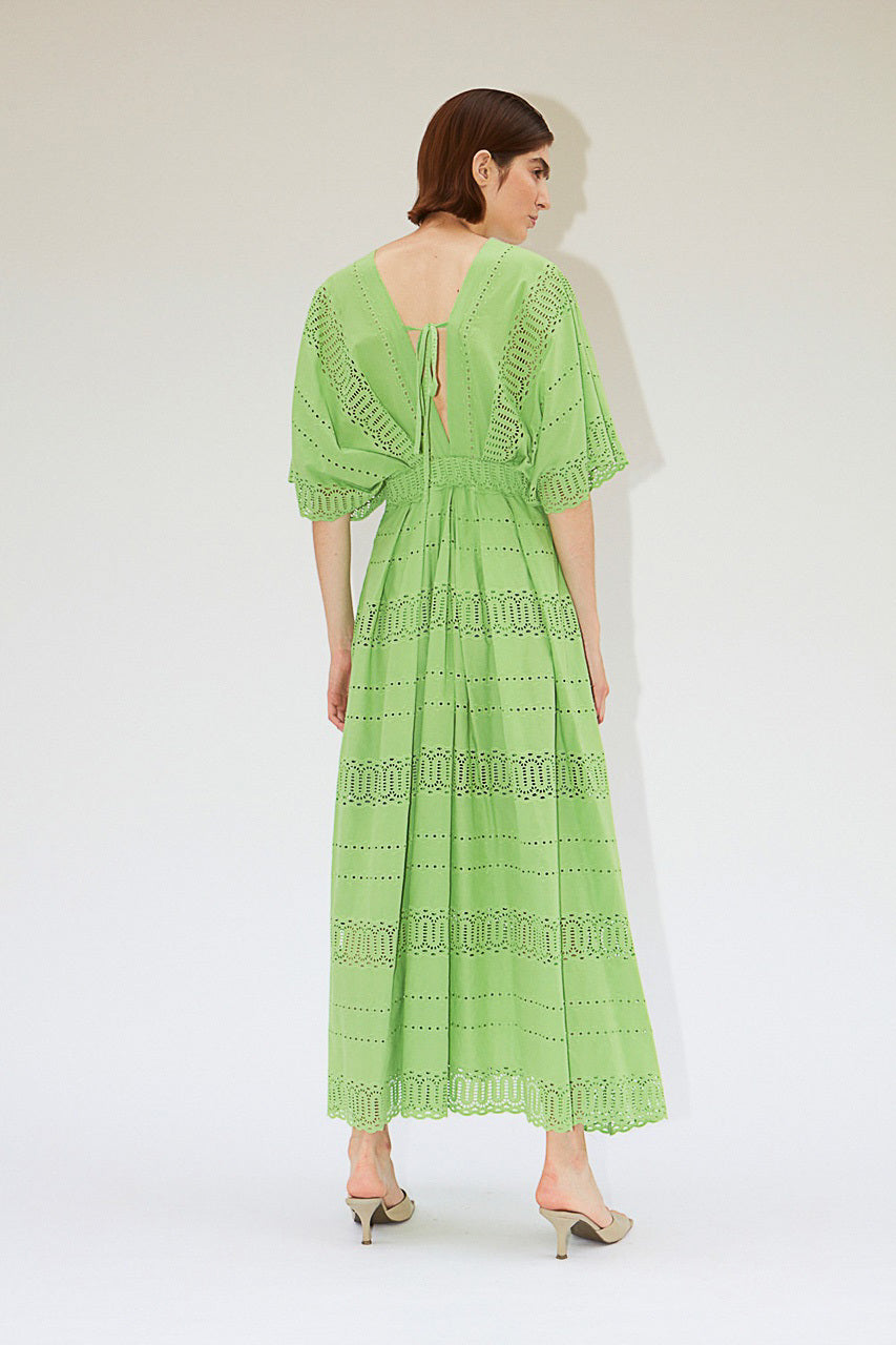 A person wearing the MILVA SANGALLO DRESS, a light green, full-length dress with a deep V-neckline and intricate eyelet lace details, perfect for a garden party. The dress features short, flared sleeves and a flowy skirt with horizontal eyelet patterns. The person is standing against a plain, light-textured background.