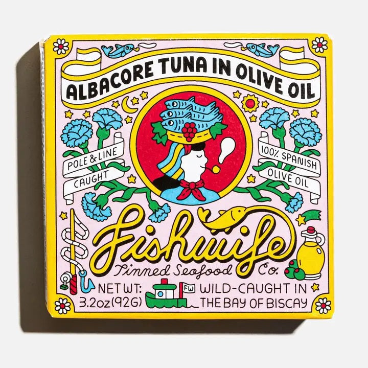 A vibrant tin of FISHWIFE Albacore Tuna in Olive Oil features a vintage-inspired design with a woman, flowers, lemons, and banners highlighting the product’s wild-caught and MSC-certified albacore tuna packed in Spanish olive oil.