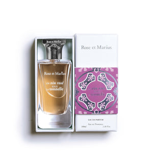A clear glass bottle of ROSE ET MARIUS - A ROSE WINE UNDER THE ARBOR Eau de Parfum 100ml, known for its fruity woody scent, with a patterned square cap beside it on a white background. The bottle casts a soft shadow.