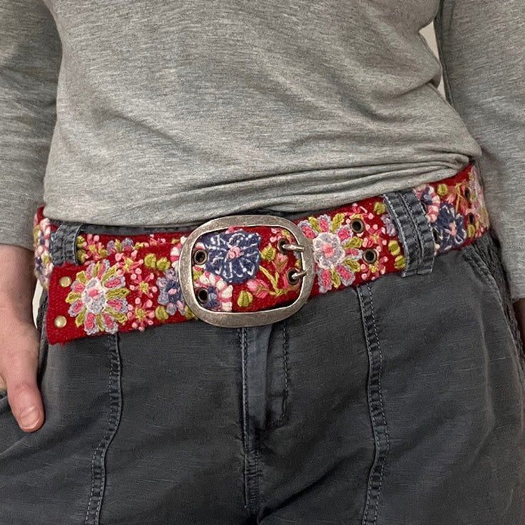 The JENNY KRAUSS - TEABERRY BELT by JENNY KRAUSS is a handcrafted, adjustable red belt with a silver buckle, featuring colorful embroidered floral patterns in pink, purple, green, blue, and white. Made in Peru.