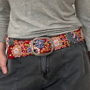 The JENNY KRAUSS - TEABERRY BELT by JENNY KRAUSS is a handcrafted, adjustable red belt with a silver buckle, featuring colorful embroidered floral patterns in pink, purple, green, blue, and white. Made in Peru.