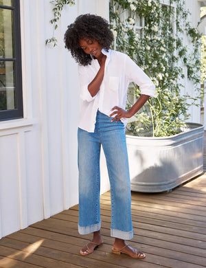 A person stands on a wooden deck with their back to the camera, wearing the FRANK & EILEEN WESTPORT Illusion Cuffed Hem Pull On Jean in 2013 Wash, styled with a white shirt and sandals. A large metal planter filled with greenery sits beside them.
