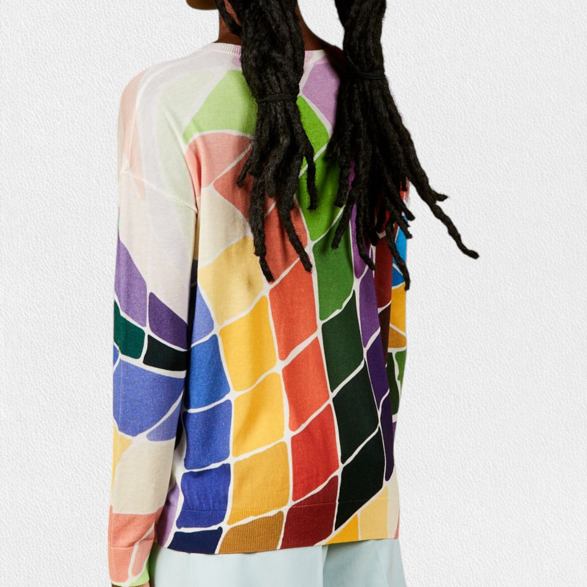 A person wearing the IN BED WITH YOU - BATWING SLEEVE V NECK JUMPER stands against a plain, light-colored background. The colorful geometric pattern featuring squares and circles in shades of pink, purple, red, yellow, and white adds flair to this stylish piece by the Italian brand IN BED WITH YOU/CLAUDIOLAI SRL. The person is visible from the shoulders down to the waist.