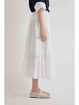 A person stands sideways in a MEIMEIJ cotton poplin midi skirt with bow detail and gathered tiers, paired with a black rolled-sleeve top and white mesh ankle-strap shoes. The background is plain and light-colored.
