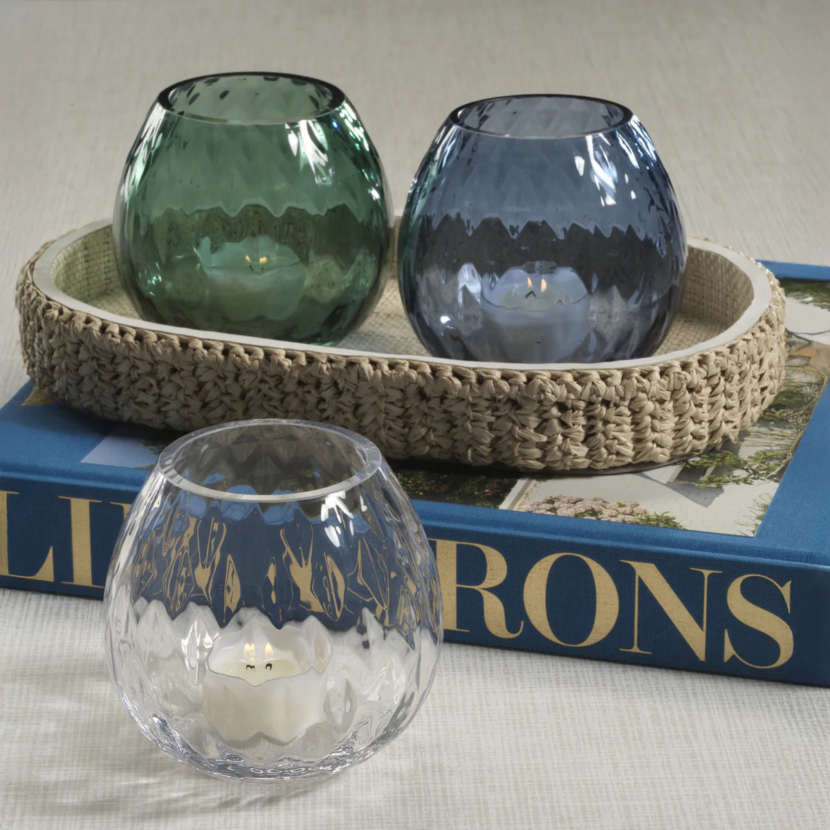 Three PRISM Multipurpose Optic Vase Candleholders by ZODAX in Deep Water Blue textured soda-lime glass, each approx. 4”, hold tealights; two in a woven tray, one on a blue book.
