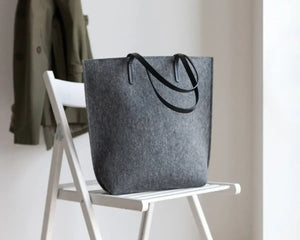 A person in a beige trench coat holds the WOOLBERRY WOOL FELT LONG TOTE BAG with black handles, standing against a plain, light-colored background.