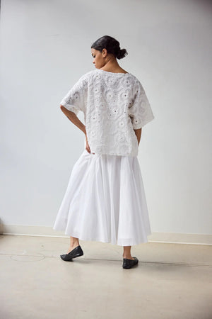 A woman stands in profile wearing the HOPE FOR FLOWERS EYELET TEE, a white, short-sleeved textured lace top made from GOTS certified organic cotton, paired with a flowing white skirt. Her hair is pulled back with a plain light background.
