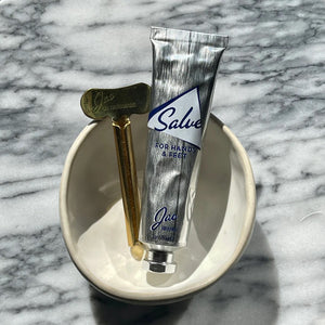 A silver tube labeled "JAO BRAND - SALVE FOR HANDS AND FEET" by JAO LIMITED, featuring essential oils, is placed inside a small white bowl. Beside the tube is a metallic golden key-like tool. The bowl sits on a marble surface.