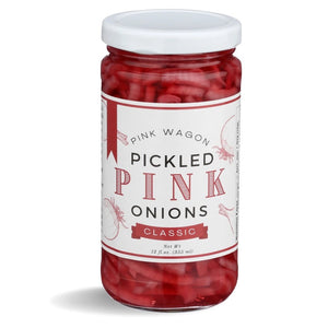 A clear jar with a white lid, labeled "PINK WAGON- PICKLED PINK ONIONS." The jar contains sliced Pacific Northwest onions in a pickling solution, filling the entire container. The label, branded with "PINK WAGON FOODS," features decorative red and white elements and indicates a net weight of 12 fl oz (355 ml).