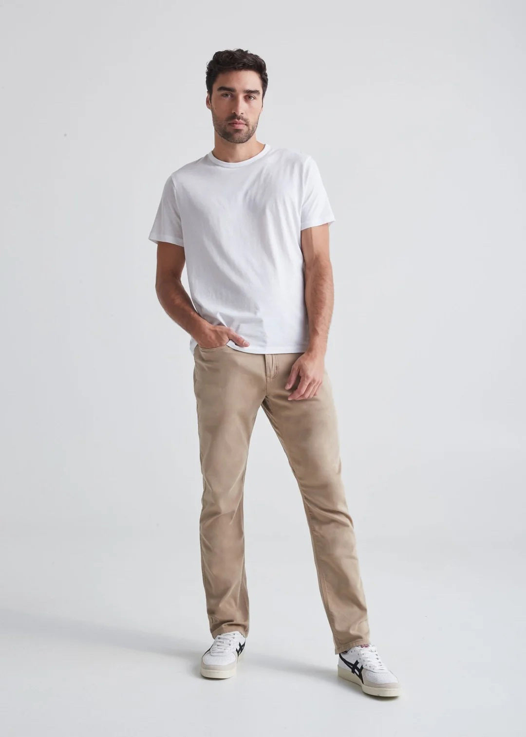 A man with short dark hair and a trimmed beard stands against a white background, wearing a white t-shirt, white sneakers with black details, and DUER's DU/ER NO SWEAT RELAXED TAPER PANT in an athletic fit. One hand is in his pocket.