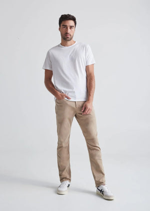 A man with short dark hair and a trimmed beard stands against a white background, wearing a white t-shirt, white sneakers with black details, and DUER's DU/ER NO SWEAT RELAXED TAPER PANT in an athletic fit. One hand is in his pocket.