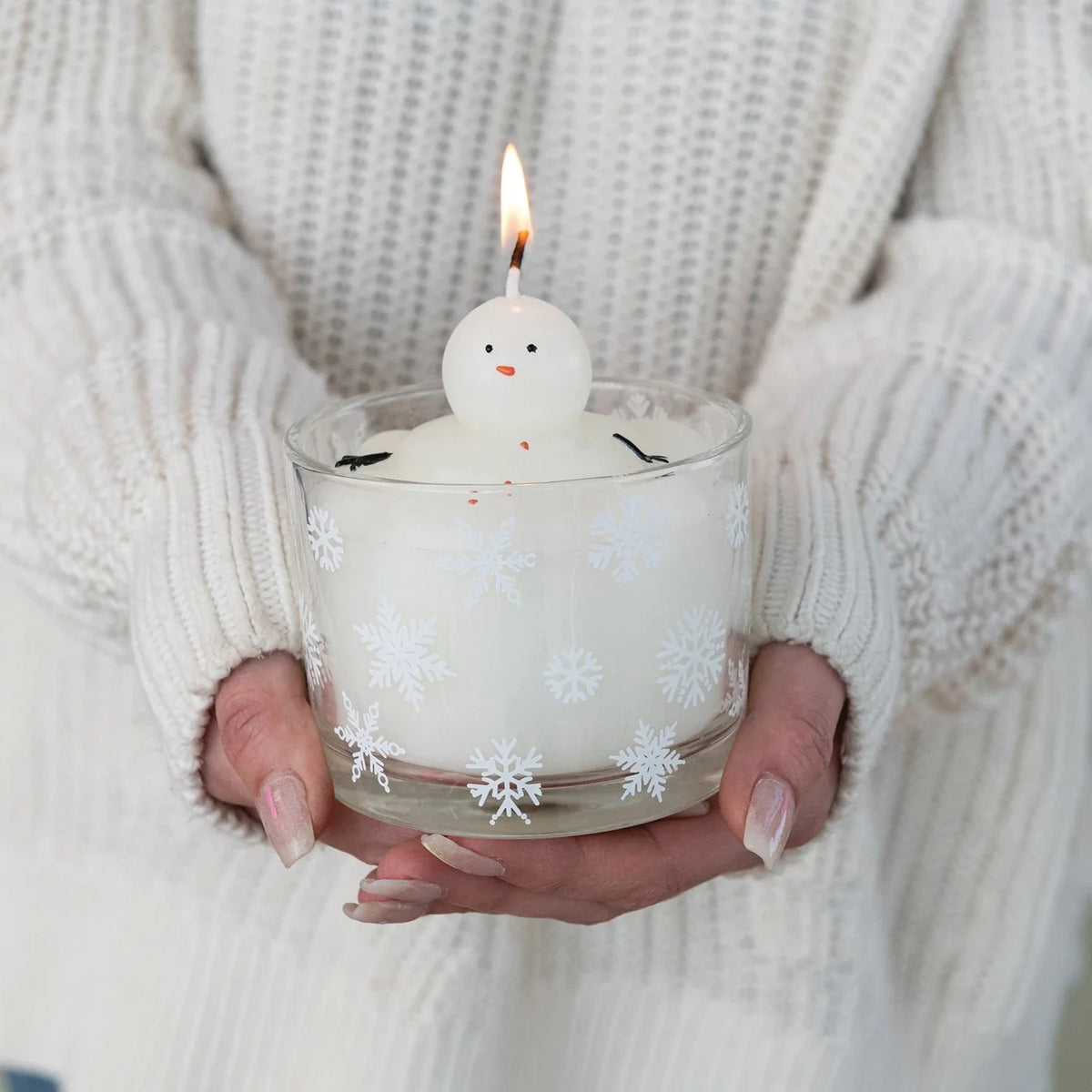 The CREATIVE COOP MELTING SNOWMAN CANDLE is a white holiday candle in a glass holder with snowflakes and a small snowman figure on top, set against a light, textured backdrop—perfect for festive winter decor.