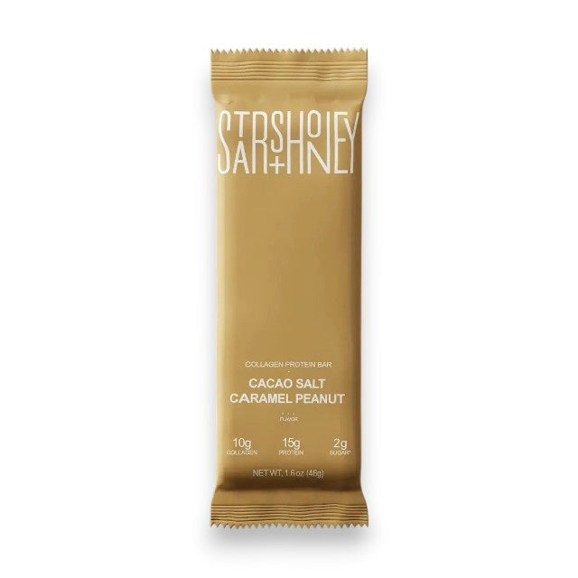 A gold protein bar wrapper labeled “STARS+HONEY” featuring the flavor “Cacao Salt Caramel Peanut” is placed on a beige background, highlighting 10g collagen, 15g protein, and just 2g sugar as a low sugar protein bar option.