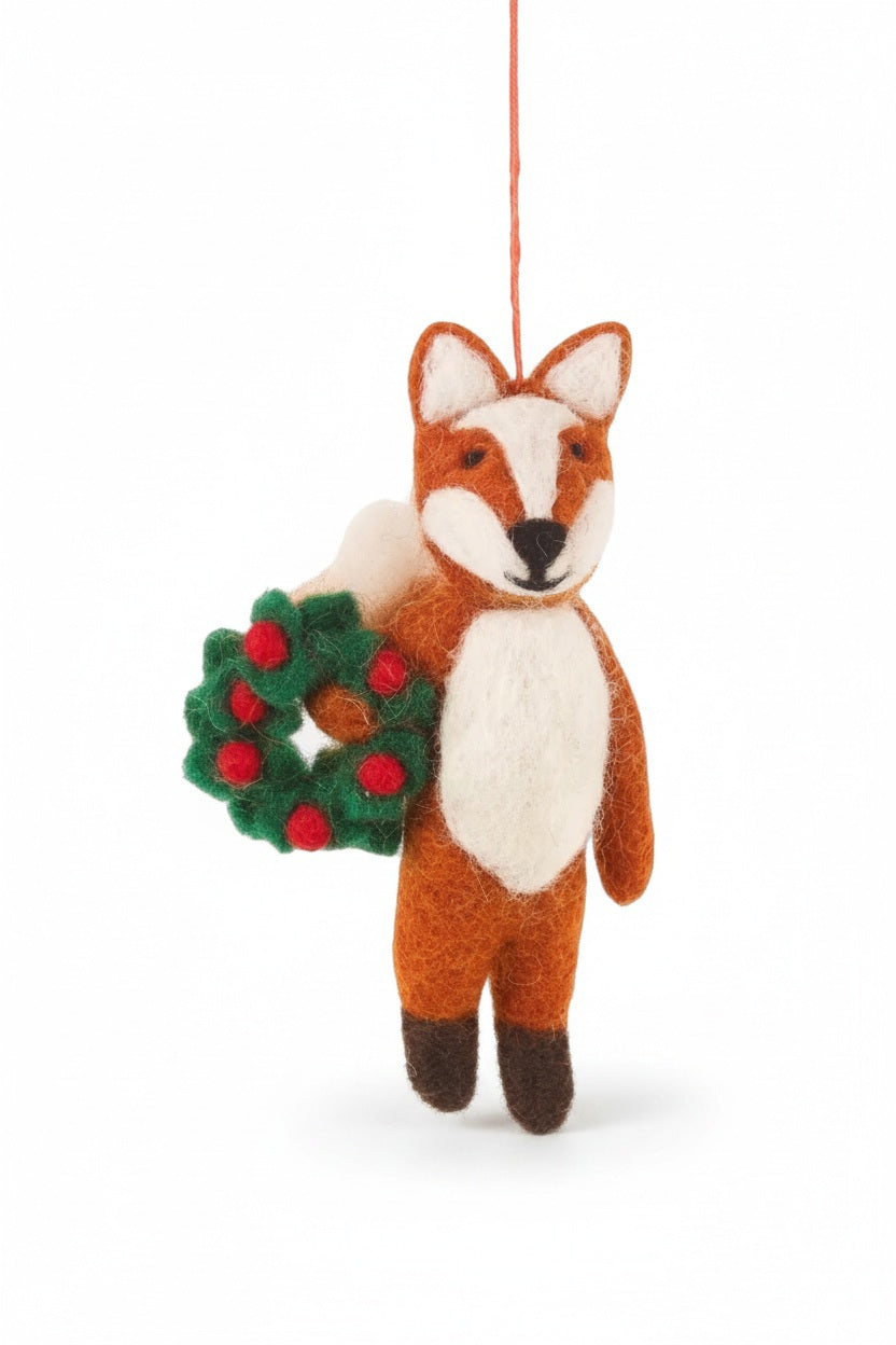 FINLEY FESTIVE FOX ORNAMENT, a handmade felt fox holding a small green wreath with red berries, standing in the snow.