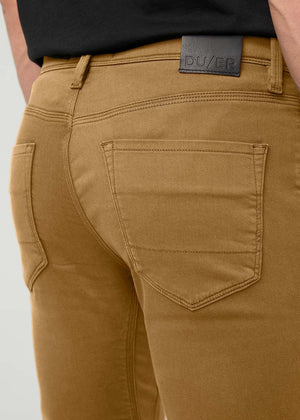 A person is shown from the lower body and part of the torso, standing against a white background while wearing DUER’s No Sweat Straight Pants in Caramel paired with a black shirt and black sneakers.