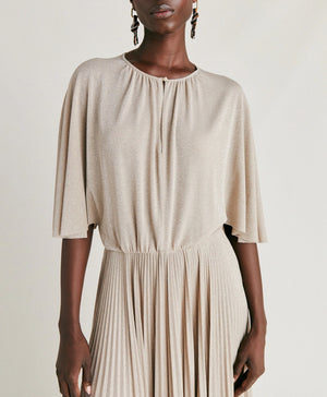 A person stands facing the camera, wearing the MOMONI - PARADISU DRESS, a light-colored, flowing pleated outfit with wide pants and kimono sleeves. Their hair is styled in neat braids, and they accessorize with drop earrings. The neutral background highlights the elegance of the MOMONI dress.